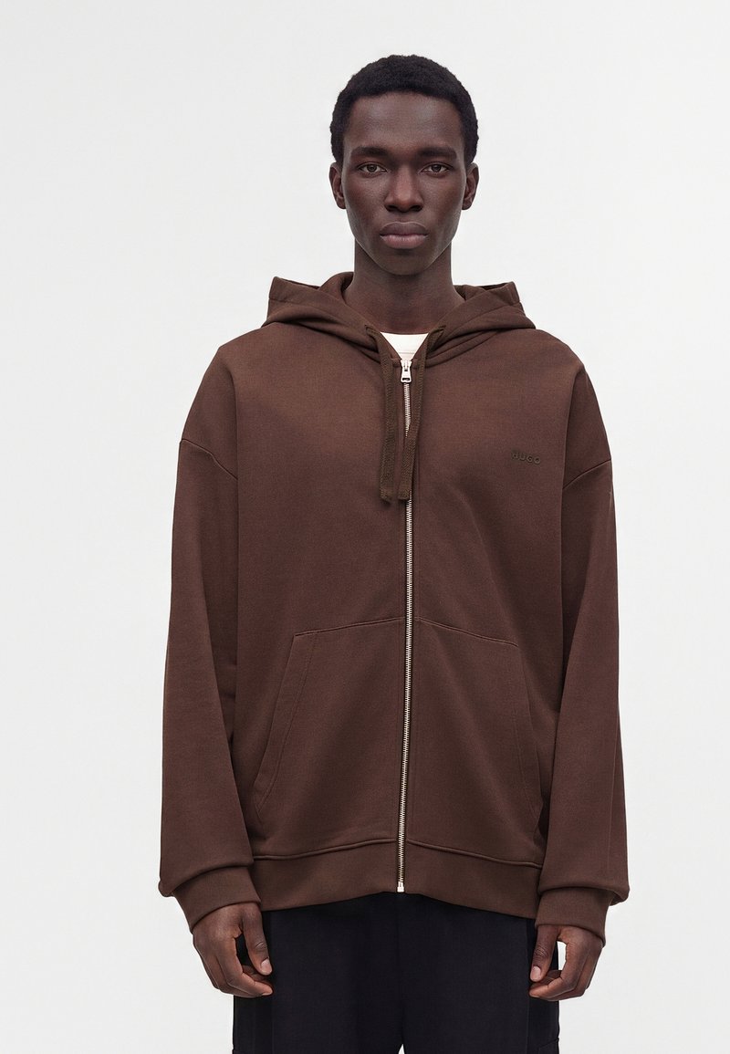 Jeune homme portant un sweat à capuche marron zippé avec des poches avant, debout devant un fond blanc uni.
