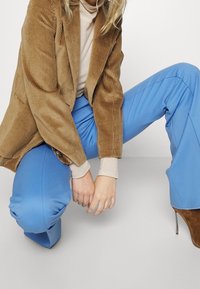 Brun fløjlskåbe, cremet turtleneck og blå skræddersyede bukser. Outfittet er suppleret med brune højhælede støvler.