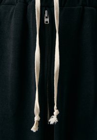 Black textured fabric joggers feature a front zipper with silver hardware and contrasting cream drawstring with knotted ends.