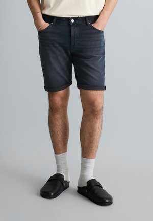 Mężczyzna w czarnych, podwiniętych szortach jeansowych, białych skarpetkach i czarnych wsuwanych butach, stoi z rękami w kieszeniach na szarym tle.