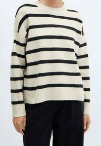 Kremowy sweter z czarnymi poziomymi paskami, okrągłym dekoltem i ribbowymi mankietami. Miękka tekstura i luźny krój, noszony z czarnymi spodniami.