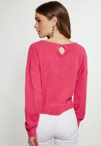 Maglione rosa soffice con scollatura rotonda, dettaglio a chiave sul retro e orlo a costine. Caratterizzato da maniche lunghe e leggermente voluminosi per una vestibilità rilassata.