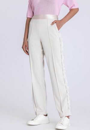 Frau trägt cremefarbene Hose mit hoher Taille, Satinbund und Seitenstreifen, kombiniert mit weißen Sneakers und einem rosa Kurzarmtop.