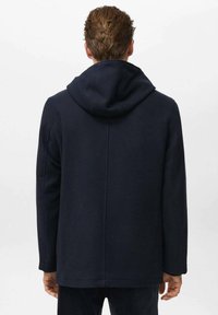 Hombre vestido con un abrigo con capucha de color azul marino oscuro, dando la espalda al espectador, frente a un fondo gris claro liso.