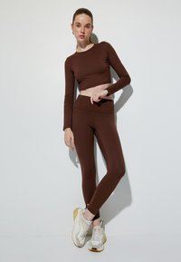 Brązowy krótki top z długim rękawem w połączeniu z pasującymi legginsami z wysokim stanem, wykonanymi z elastycznego materiału. Model nosi beżowe tenisówki. Prosty, elegancki fason.