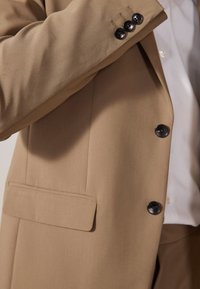 Chaqueta de traje color marrón claro con una textura suave, tres botones negros en el puño y un bolsillo. Debajo lleva una camisa blanca.