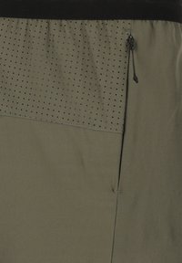 Olivgrön tyg med en strukturerad panel som har små svarta prickar. Inkluderar en svart elastisk midjeband och en blixtlåsdetalj på sidan.
