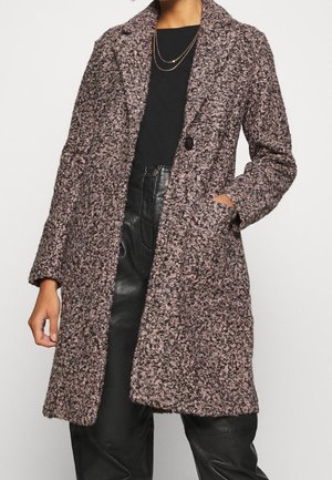 Long manteau texturé rose et noir sur un top noir et un pantalon en cuir noir taille haute, main dans la poche, colliers en or superposés.