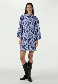 Vestido camiseiro azul e branco com padrão, com fecho de botão na frente, mangas bufantes longas e corte reto. Usado com botas pretas até ao joelho.