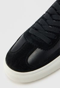 Μαύρο αθλητικό παπούτσι που συνδυάζει σουέντ και λείο δέρμα, με στρογγυλεμένη μύτη, λευκή σόλα από καουτσούκ και μαύρα κορδόνια με υφή.