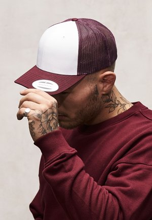 Rode en witte snapback pet met een mesh achterkant, platte rand en een ronde label aan de voorkant. De persoon draagt een bordeauxrode sweatshirt en rookt.