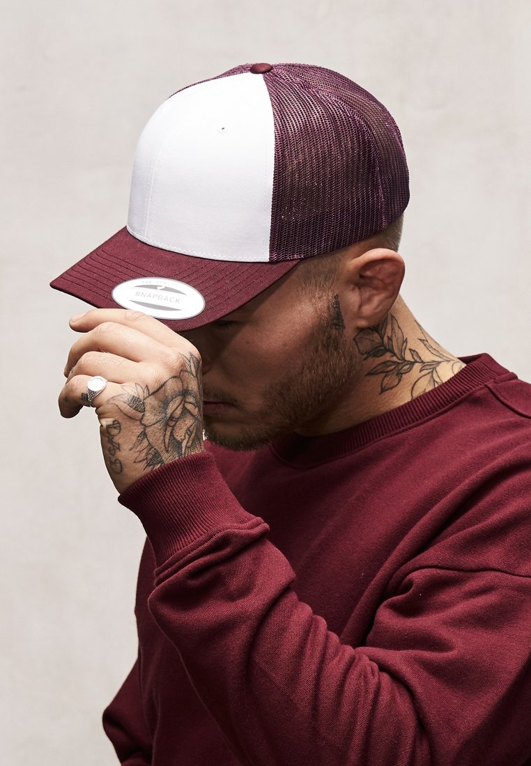 Crvena i bijela snapback kapa s mrežastim strahom, ravnim vizirima i kružnom etiketom na prednjem dijelu. Osoba nosi bordo duksericu i puši.
