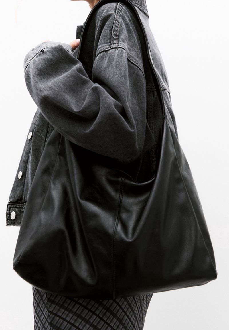 Personne portant une veste en jean foncée tient un grand sac à bandoulière en cuir noir devant un fond clair uni.