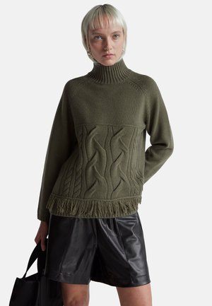 Pull en tricot vert olive avec un col haut, motif en câble et finitions à franges. Associé à des short en cuir noir.
