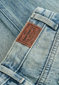 Heled sinine denimkangas tekstureeritud pinnaga; sisaldab pruuni nahkset sildiret, millel on tumedates tähtedes sissepressitud "DEAN GOODS CO".