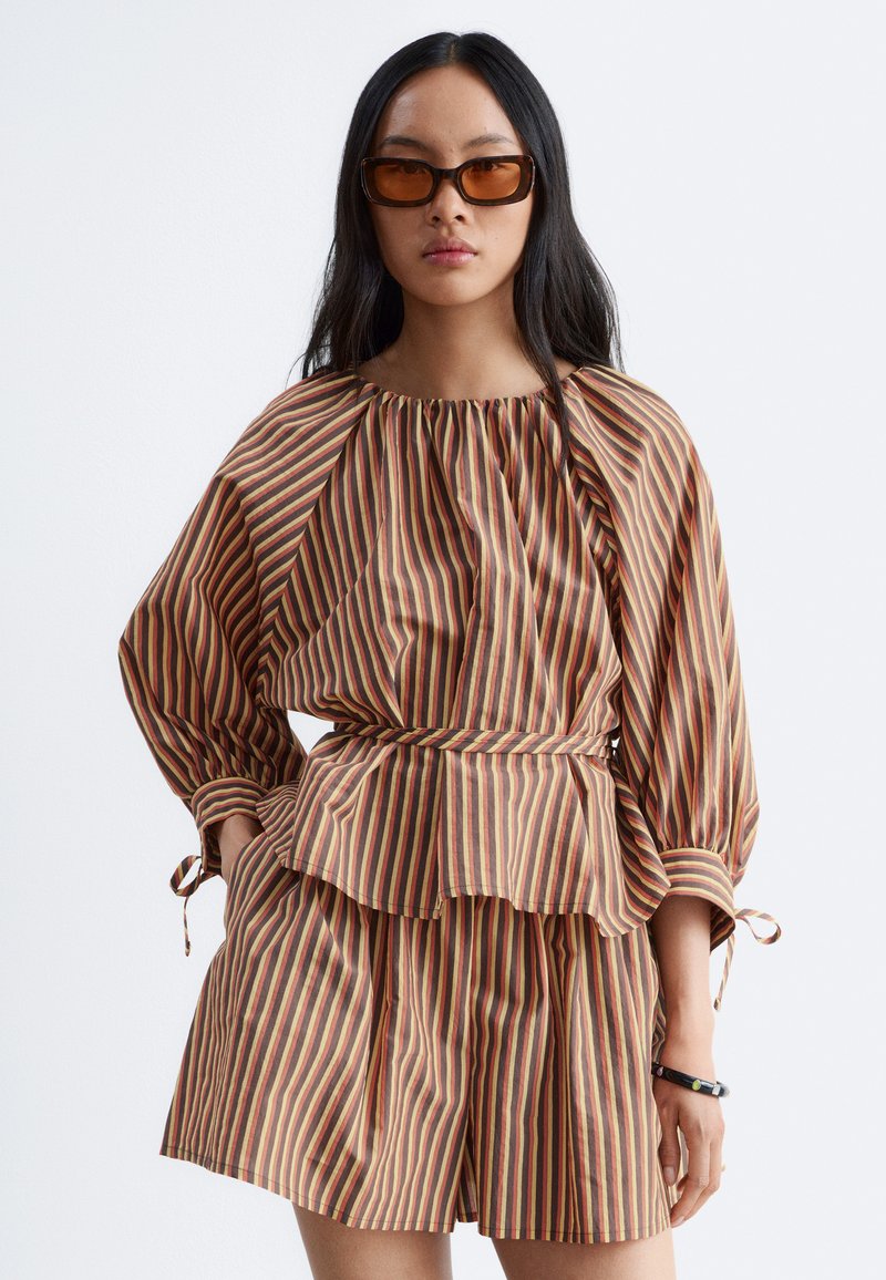 Femme portant un haut à manches longues à rayures verticales marron, rouge et beige et un short assorti, avec des lunettes de soleil marron et un bracelet noir.