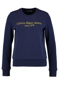Námořnická modrá mikina vyrobená z bavlny, s žebrovanými manžetami a spodním lemem. Na přední straně je vyšitý nápis "Calvin Klein Jeans since 1978" v jasně žluté barvě.
