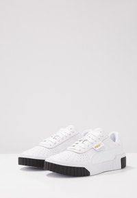 Zapatillas blancas de cuero con textura perforada, suela plana de goma negra, cordones blancos y logo dorado de Puma en el costado. Diseño elegante.