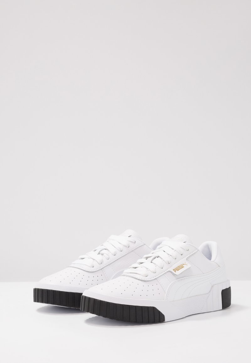 Baskets en cuir blanc avec texture perforée, semelle en caoutchouc noir plate, lacets blancs et logo Puma doré sur le côté. Design élégant.