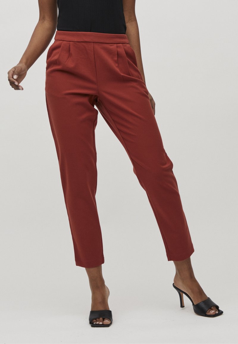Femme portant un pantalon fuselé taille haute couleur rouille et des mules à talons ouvertes noires, debout devant un fond clair uni.