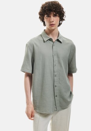 Camisa de botón de manga corta de tela verde claro, con cuello, acabado texturizado y bolsillos frontales. Corte simple y relajado.