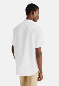 Camicia bianca di lino a maniche corte con colletto classico, caratterizzata da un orlo curvo e una texture liscia; dettaglio di cucitura sottile sulle spalle.