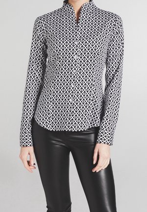 Femme portant une chemise boutonnée à motifs géométriques noir et blanc avec col mao et un pantalon en cuir noir, debout devant un fond gris.