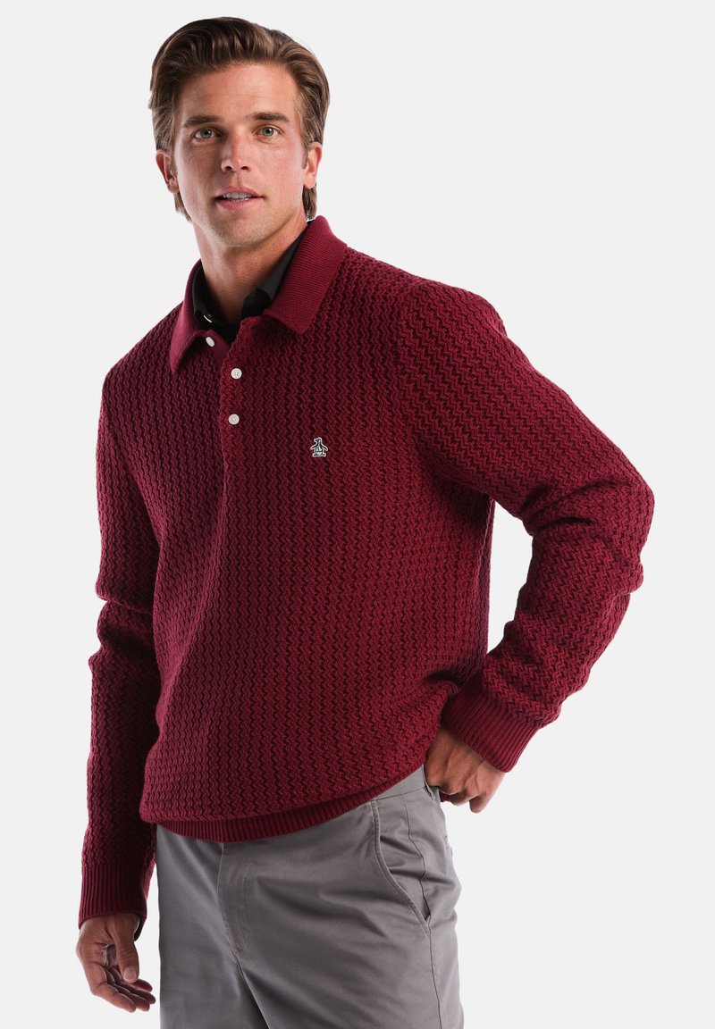 Bordeauxfarbener Strickpullover mit einem strukturierten Muster, einem gerippten Kragen, drei Knöpfen und einem kleinen Logo auf der Brust. Getragen mit grauen Hosen.