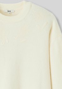 Pull en maille couleur crème avec motifs floraux en relief près de l'encolure ronde et une étiquette taille M à l'intérieur du col.