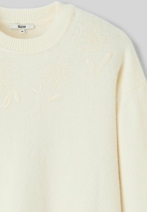Pull en maille couleur crème avec motifs floraux en relief près de l'encolure ronde et une étiquette taille M à l'intérieur du col.