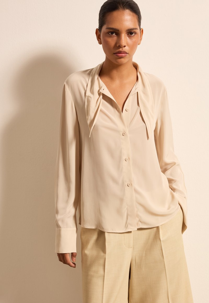 Beige Bluse aus einem weichen, seidigen Material mit einem gebundenen Kragen, Knopfleiste und langen Ärmeln, kombiniert mit hellbeigen Hosen.