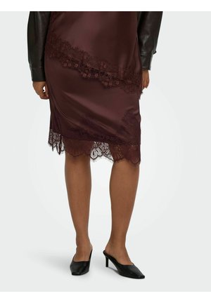 Femme portant une jupe en satin marron foncé avec bordure en dentelle et une veste en cuir noire, debout dans des mules à talons noires sur fond blanc.