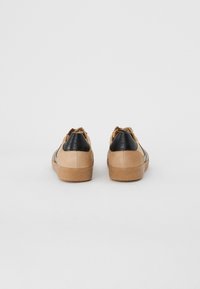 Sneaker in suede tan con accenti in pelle nera, suole in gomma texture e strisce a contrasto. La vista posteriore mostra un design minimale.