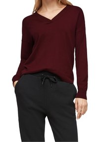 Frau trägt einen burgunderroten V-Ausschnitt-Pullover mit langen Ärmeln und schwarze Hose mit Kordelzug, Hände in den Taschen.