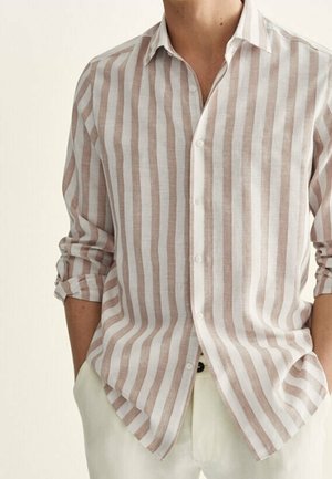 Man draagt een beige-witte overhemd met verticale strepen, met de mouwen opgerold tot de ellebogen en handen in de zakken van een witte broek.