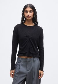 Μαύρο μακρυμάνικο τοπ με υφή υφάσματος, λαιμόκοψη και τελείωμα με κυματιστό σχέδιο, που διαθέτει κούμπωμα με κουμπιά στο μπροστινό μέρος. Συνδυασμένο με γκρι παντελόνια.