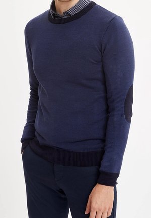 Homme portant un pull texturé bleu marine avec des coudières noires et une chemise à carreaux, les mains dans les poches, sur fond blanc.