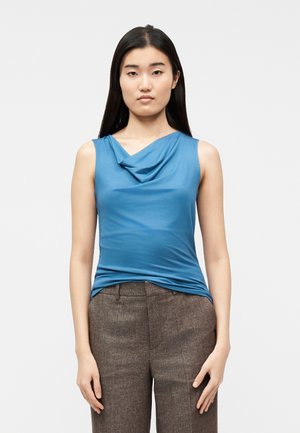 Jeune femme aux longs cheveux foncés portant un haut bleu sans manches à encolure drapée et un pantalon marron ajusté, debout devant un arrière-plan uni.