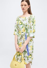 Femme portant une robe blanche à motifs de citrons et de feuilles vertes, tenant un sac à main jaune, debout devant un fond clair uni.