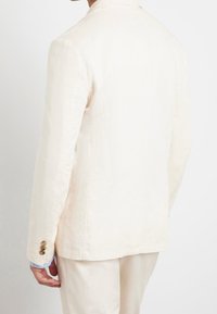 Světlý béžový blazer, přiléhavý střih se dvěma knoflíky na každém rukávu, hladká textura, mírně strukturovaná látka a klopy ve tvaru písmene "notch".