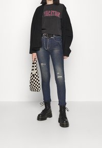 Osoba ubrana w czarny kardigan, ciemnoszary T-shirt z napisem "VACATION", przetarte skinny jeans, czarne platformy oraz trzymająca torbę w czarno-białą kratę.