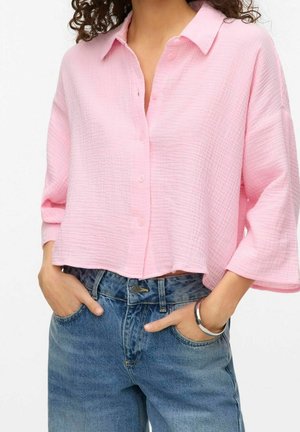 Mujer con blusa holgada de botones de color rosa claro y tela texturizada, jeans azules de cintura alta, con las manos metidas en los bolsillos y una pulsera plateada en la muñeca.