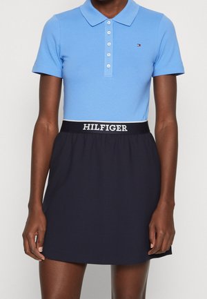 Vrouw draagt een lichtblauw poloshirt met korte mouwen en een zwarte rok met een tailleband waarop het merk "HILFIGER" staat.