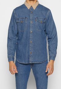 Uomo che indossa una camicia di denim blu con colletto beige e jeans blu abbinati, in piedi davanti a uno sfondo bianco uniforme.