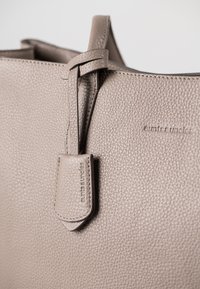 Graue Lederhandtasche mit einer strukturierten Oberfläche, einem schlanken Griff und einem rechteckigen Anhänger, auf dem der Markenname eingeprägt ist.