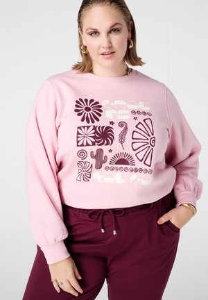 Sweat-shirt rose clair avec des motifs graphiques audacieux en violet foncé, comprenant des fleurs et des formes, associé à un pantalon bordeaux avec une ceinture réglable.
