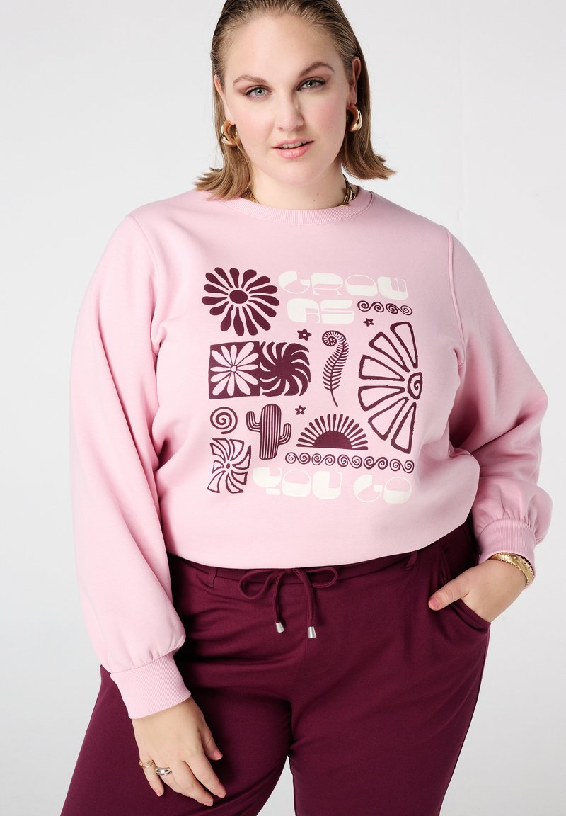Lichtroze sweatshirt met gedurfde donkerpaarse grafische ontwerpen, waaronder bloemen en vormen, gecombineerd met bordeauxrode broek met verstelbare tailleband.