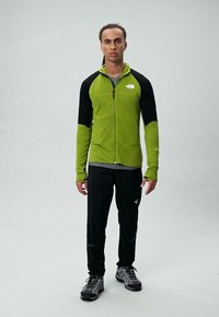 Męski strój sportowy składający się z zielonej polarowej kurtki z czarnymi rękawami, czarnych spodni i szarych butów turystycznych, idealny do aktywności na świeżym powietrzu.