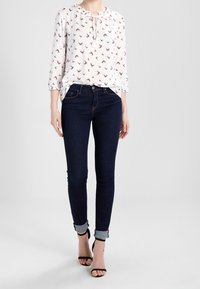 White blouse with red bird print, keyhole neckline, and three-quarter sleeves paired with dark blue skinny jeans and black heeled sandals.