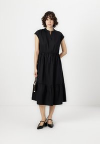 Black, knee-length dress with a gathered waist, short sleeves, and a buttoned neckline. Model holding a small black bag, wearing black shoes.